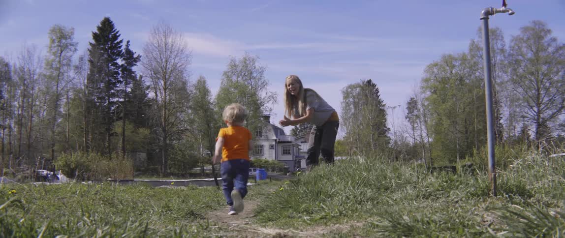 A woman claps and cheers while a young toddler boy runs past her outdoors.