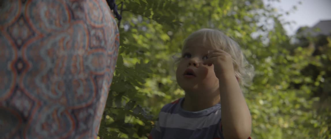 A young toddler boy stands outdoors in nature while playfully twisting his curly hair.