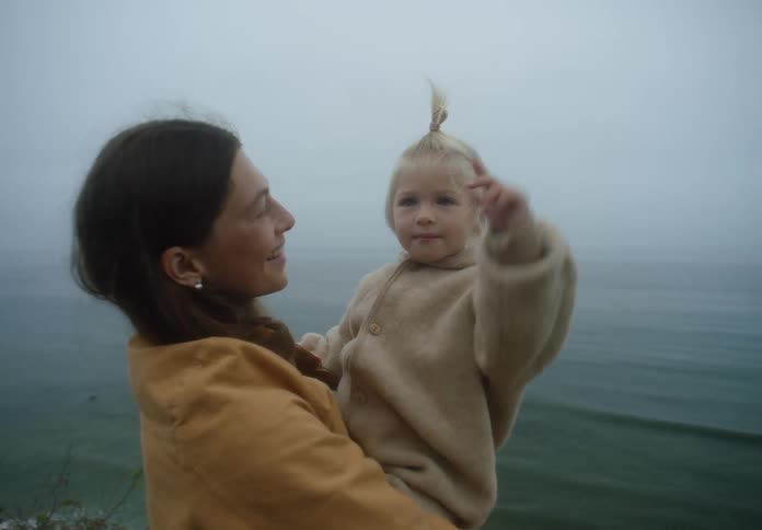 A smiling woman in a yellow coat holds a young child in a fleece jacket on a cliff overlooking the ocean during a foggy day