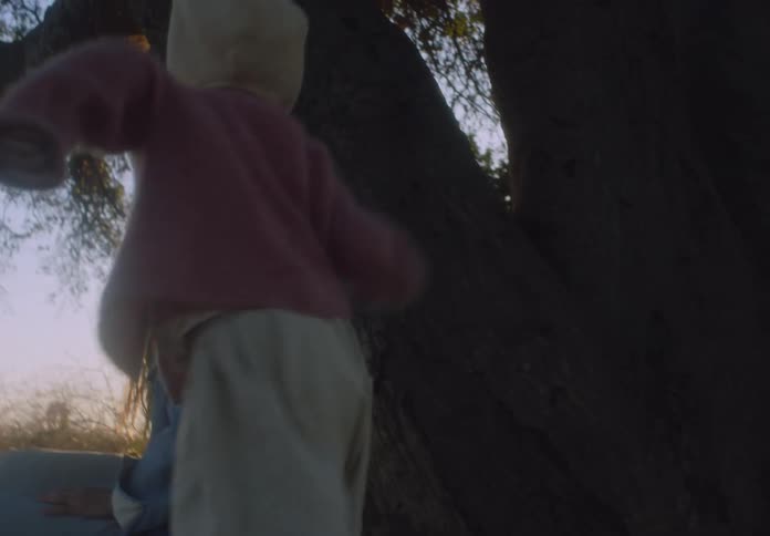 A young girl walks around a tree and then sits down on the lap of a woman