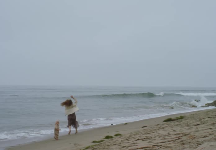A woman in a long skirt and sweater dances and spins on a foggy beach while a small toddler in a tan romper runs beside her near the crashing ocean waves under a grey overcast sky