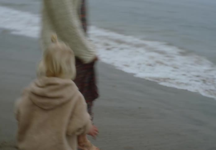 A young child in a tan hooded outfit runs toward the ocean waves on a sandy beach before a woman in a white sweater catches them during a cloudy day