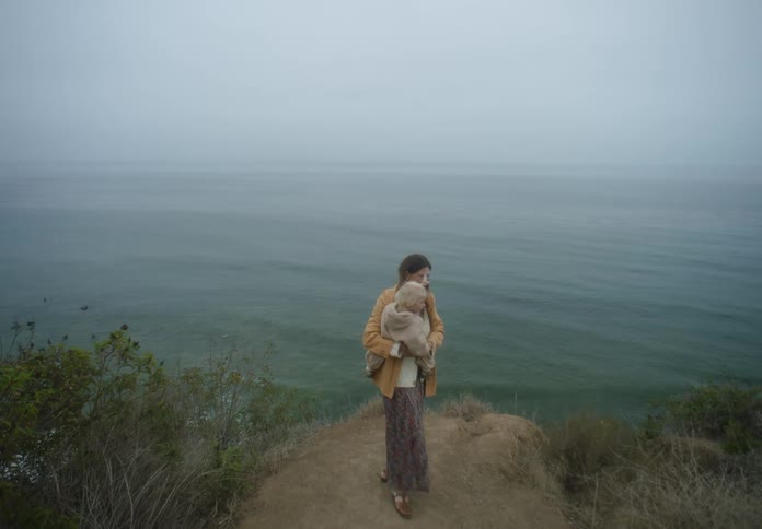 A woman wearing a yellow jacket and patterned skirt stands on a dirt cliff holding a young child in a fleece outfit while looking out over the calm ocean on a gray and misty day