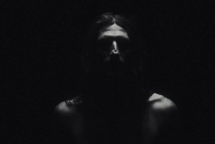 A shadowed man under flashing light with long hair is standing against a black background their face partially illuminated by a narrow beam of light