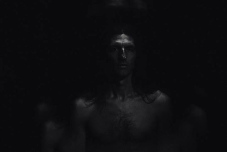 A shirtless man with long hair and a beard stares directly into the camera against a black background