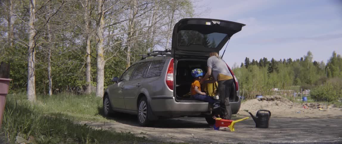 A woman puts a bike helmet on a child sitting in the open hatchback