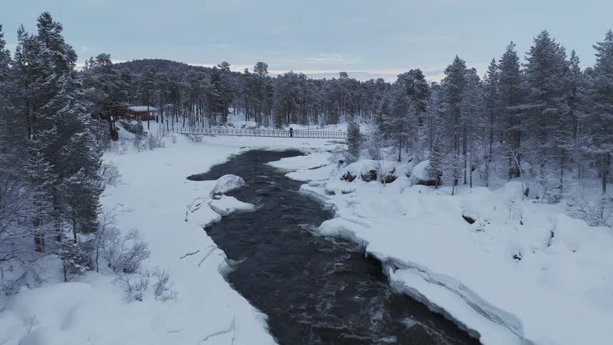 A river flowing through an arctic forest with a bridge crossing over it and someone standing on the bridge at midday