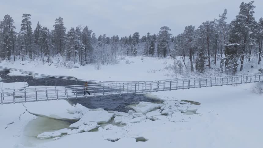 A person wearing an orange jacket walks across a long suspension bridge over a dark river flowing through a snowy forest under a gray winter sky