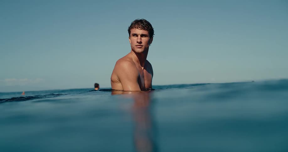 A shirtless male surfer sits on his board waiting for a wave