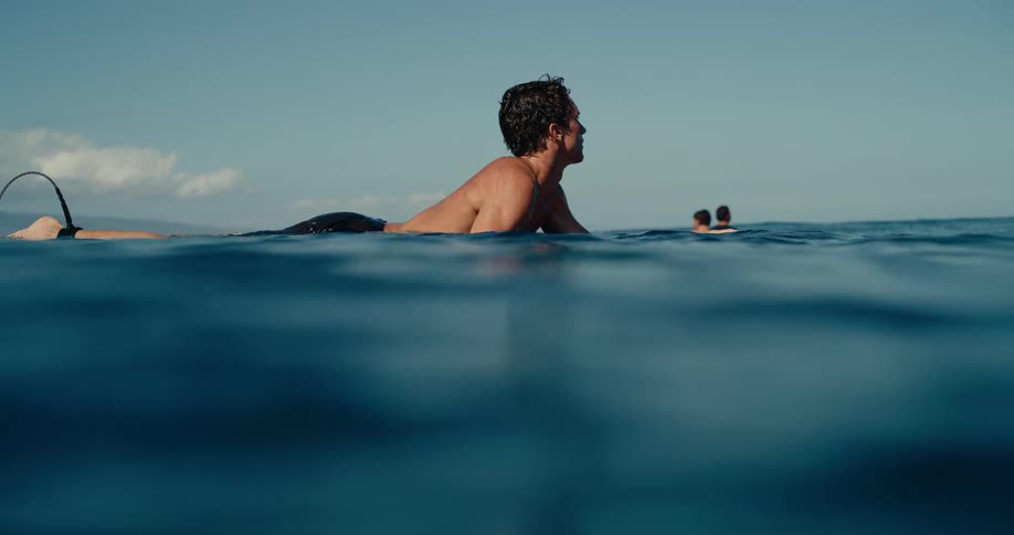 A man emerges from the dark blue water while holding a surfboard and looks around at other people in the distance under a clear sky
