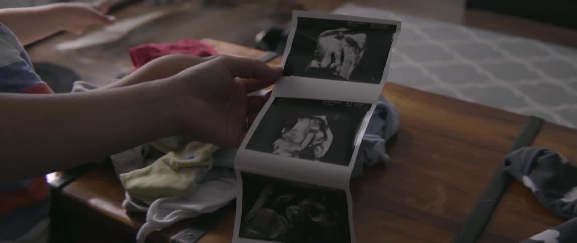 A pregnant woman holds a strip of sonogram photographs while sitting in a room.