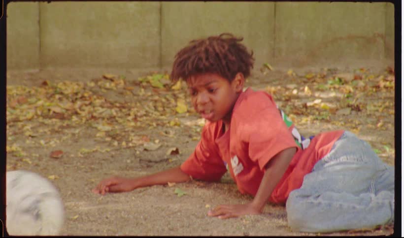 Children run through a park while kicking a soccer ball as one child lays on the ground surrounded by trees and greenery with sunlight filtering through the leaves