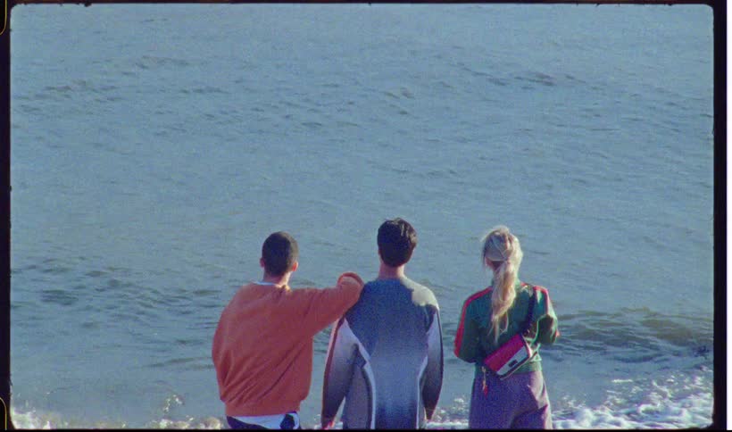 A view from behind of two men and one woman standing on a sandy beach and looking out at the blue ocean waves under a clear sky
