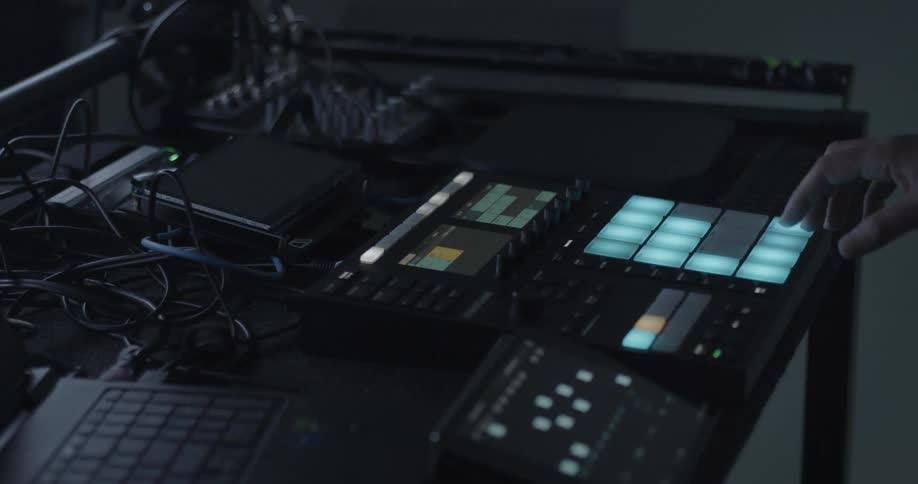 A man uses his right hand to press illuminated teal pads on a sampler while working in a dark music studio surrounded by various electronic equipment and tangled cables on a workspace