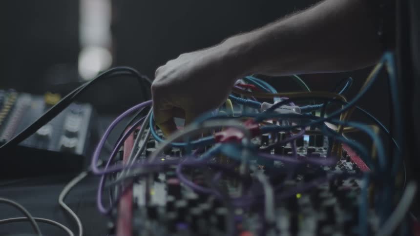 A man connects a green patch cable into a complex modular synthesizer filled with colorful wires and numerous control knobs while a professional audio mixer sits blurred in the dark background of the music studio