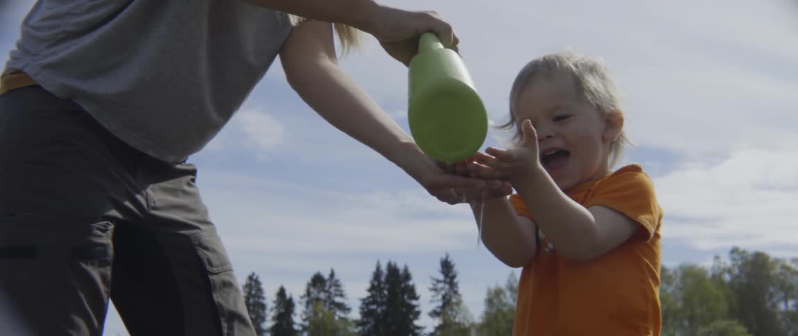 A woman washes a toddler's hands with a watering can in a sunny garden.