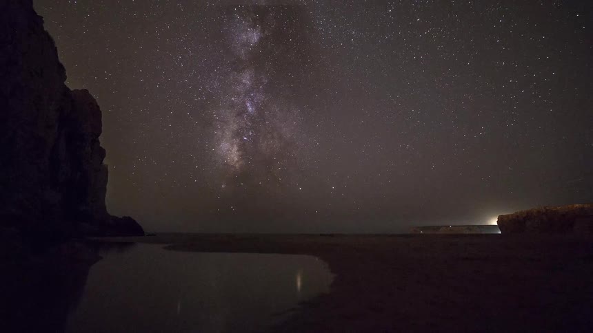 Milky way visible in the night sky over a beach with cliffs and reflection in a water pool