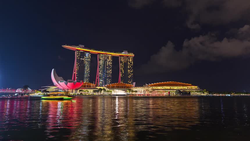 The marina bay sands hotel in singapore glows with blue and pink lights at night while a tour boat passes by on the dark water of the bay under a cloudy sky