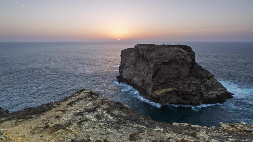 A large rock sitting in the ocean near the coastline with waves crashing against its sides and the orange setting sun in the distance as evening turns into night