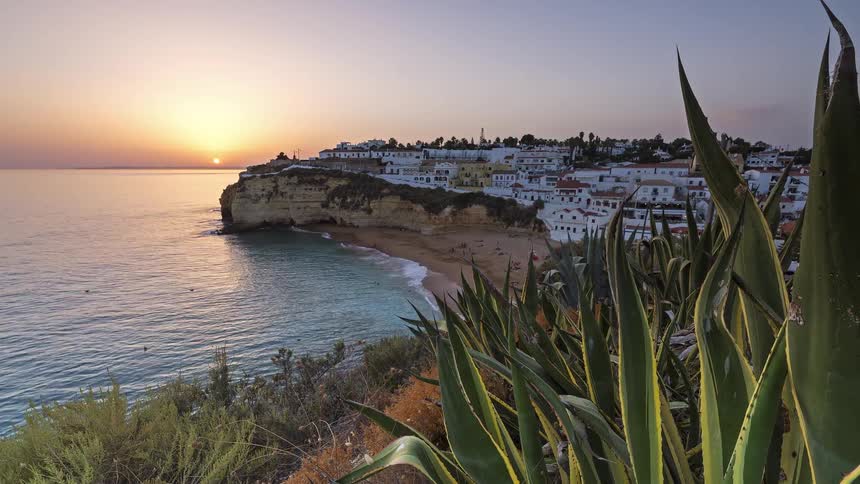 A coastal town sits atop a cliff overlooking a beach and calm ocean as the sun sets on the horizon