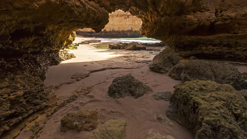Rock cave on the coast with an opening facing a sandy area scattered rocks and the sea in the background
