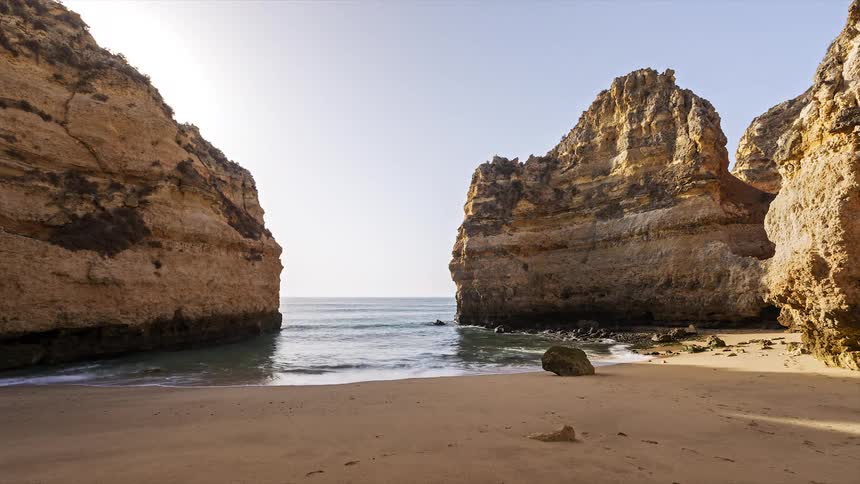 Rock cliffs overlook a woman who lays on the beach and then walks away as waves roll into shore and over the sand