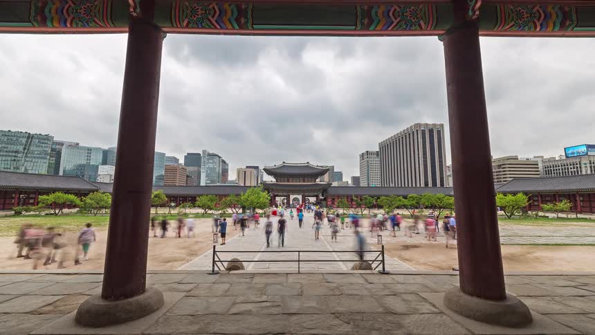 A view through the stone pillars of gyeongbokgung palace shows a large gate and people walking across a wide courtyard while modern office buildings and a grey sky fill the background in seoul