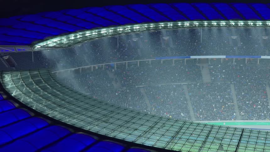 A view from above showing a large sports stadium filled with a crowd of people as rain falls under bright lights and a blue roof structure