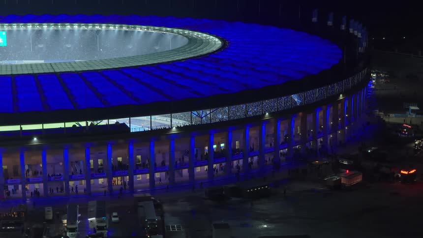 People either sit in the stands or move about the corridors of a brightly lit stadium at night