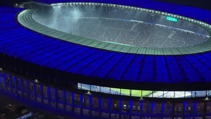 A high view looking down at a large sports stadium at night with a bright blue roof and thousands of people in the stands while white smoke drifts across the field during a game