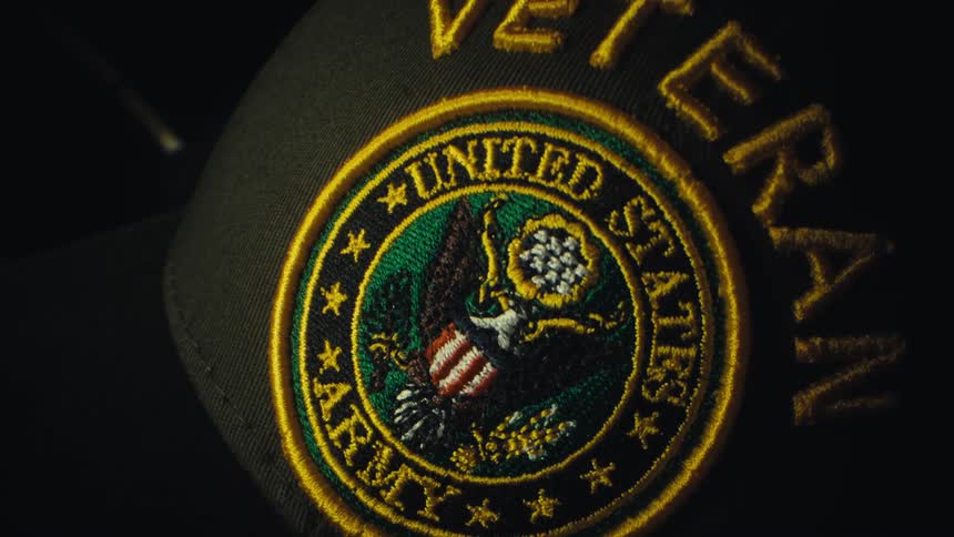 A close up shot zooms into a dark green baseball cap featuring a golden embroidered united states army veteran patch with an eagle crest and red white and blue shield against a black background