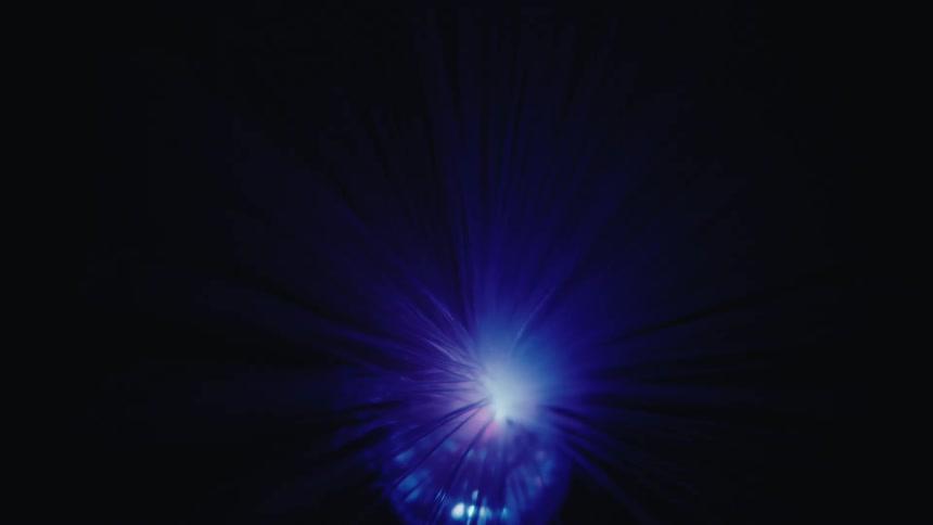 A close view of a glowing bundle of optical fibers that change from green to blue before fading into many tiny colorful dots of light against a black background