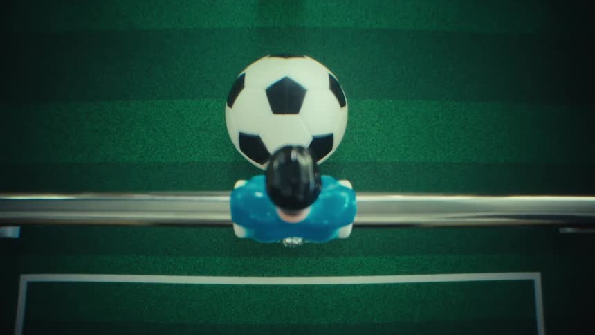 A view from above shows a blue plastic figure on a metal rod hitting a small soccer ball across a green table while playing a game of table football