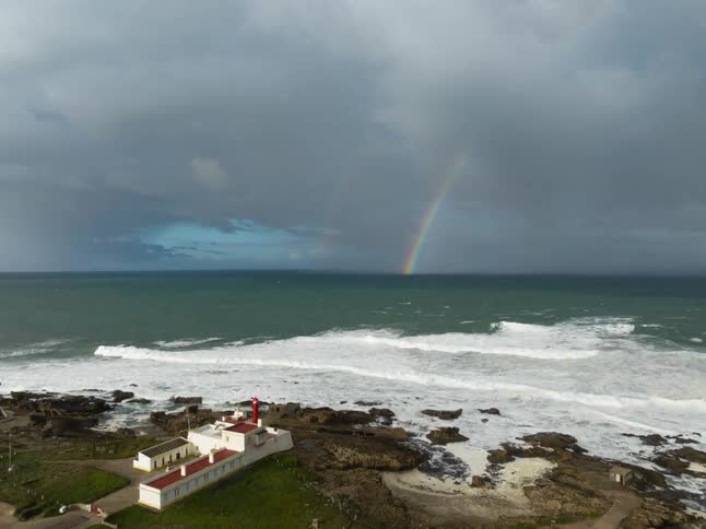 A white lighthouse with a red roof on a rocky coast under a dark stormy sky where a double rainbow fades above the white crests of a rough green ocean as waves break against the jagged shoreline stones
