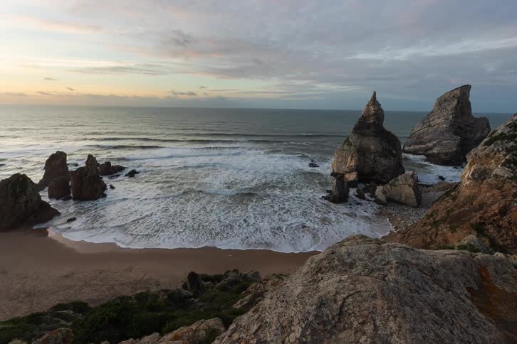 A view from a rocky cliff looks down at waves crashing onto a sandy beach with large rock formations standing in the water as the sun sets over the ocean under a soft orange and purple sky