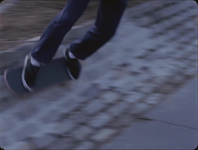 A person wearing dark pants and black shoes rides a skateboard across a slanted stone surface next to a sidewalk during the day