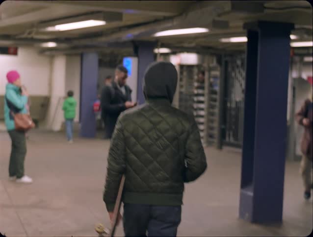 Person wearing a hood holding a skateboard walking inside a subway station
