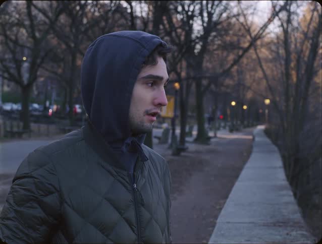 Man wearing a hood and padded jacket standing beside a paved path in a park with leafless trees