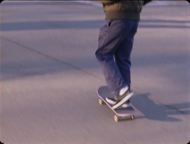 A skateboarder in a dark quilted jacket and blue pants glides down a paved park path lined with trees and benches the person kicks off to gain speed and continues riding away from the camera toward the horizon