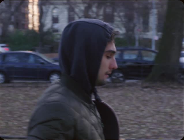 Young man walks through a park carrying his skateboard and a drinking from a cup