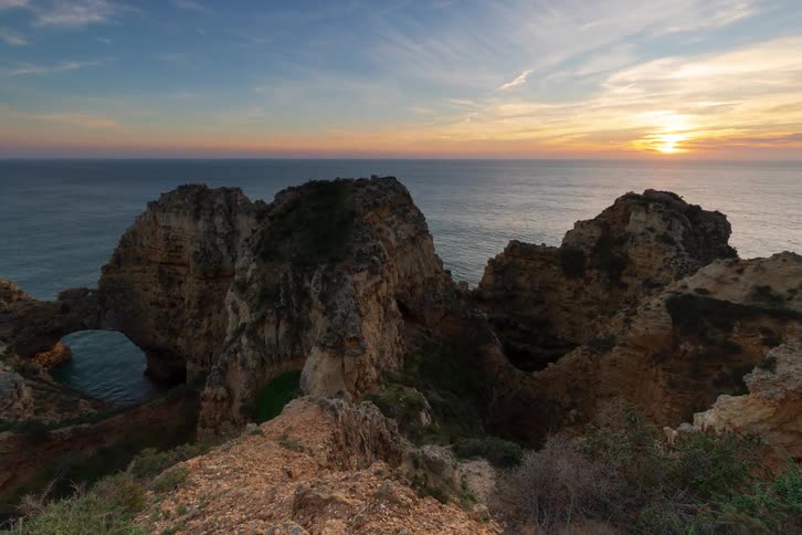 A view from a rocky cliff looks down at waves crashing onto a dark sea with large rock formations and an archway under a sunset sky that fades from orange to deep blue