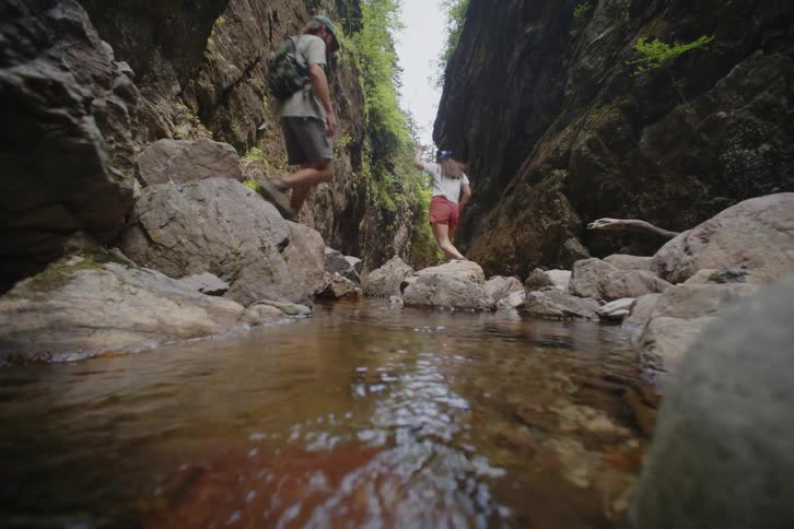 Hikers crossing a shallow stream between rocky canyon walls with vegetation