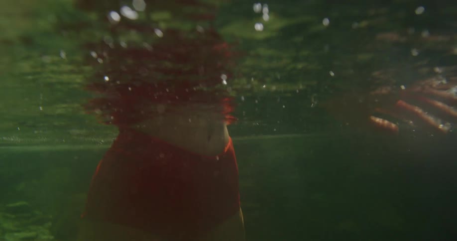 A young woman in a red and patterned bikini swims in a clear forest river on a sunny day