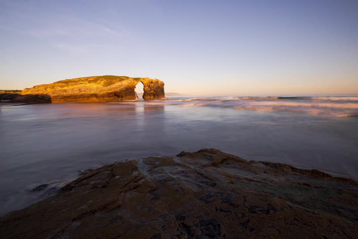Natural arch in a sunlit rock formation beside the sea with gentle waves along the shore