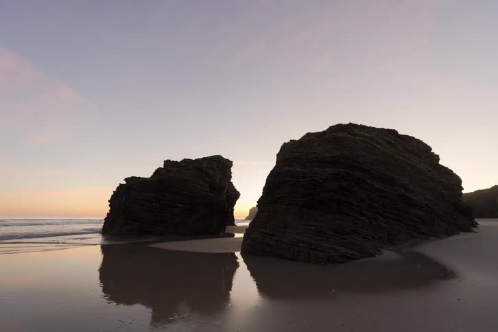Large sea stacks stand on a sandy beach with ocean waves and reflections in wet sand at sunset