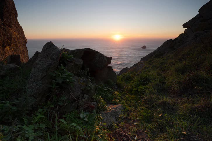 The sun sets over the ocean and a small rock island as seen from a grassy hill with large dark rocks in the foreground under a clear sky