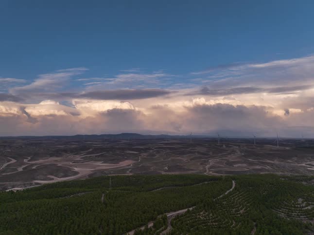 Curved crop rows in a valley with dirt roads hills and wind turbines beneath broad clouds on the horizon
