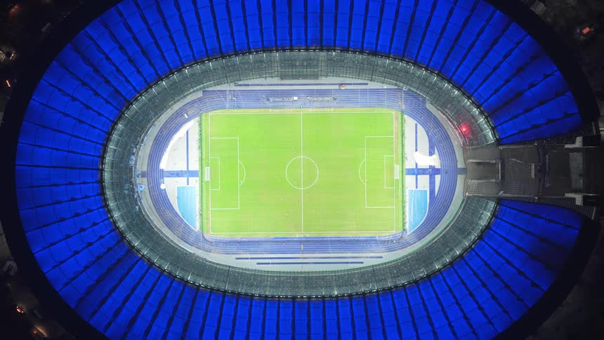A view from directly above looking down at a green soccer field inside a large stadium with a bright blue oval roof and glowing lights at night