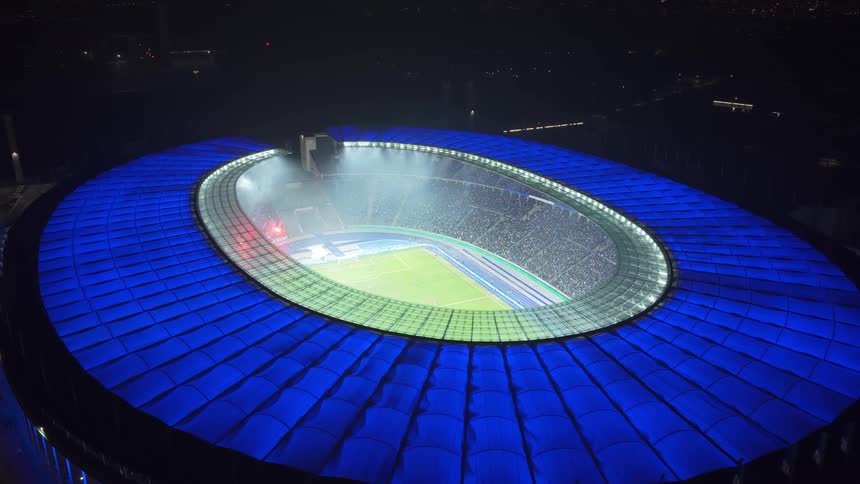 An aerial view from directly above looks down at a green soccer field inside a large stadium with a bright blue oval roof and glowing lights at night