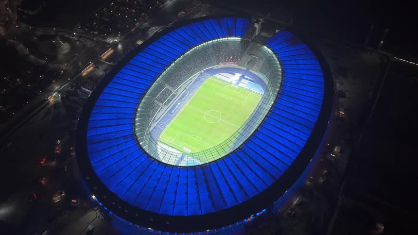A football stadium with a round blue roof lit up at night filled with football fans in the middle of the city